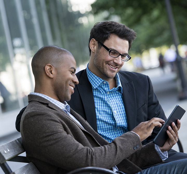 Two men in blazers on side walk bench looking at a tablet.