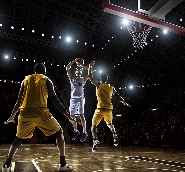 Image from a low angle viewpoint of a basketball player going to shoot a ball in a hoop  basketball in front of rival players. The action takes place in generic indoor floodlit basketball arena full of spectators. All players are wearing generic unbranded basketball uniform.