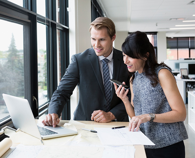 Two business people collaborating in front of a laptop while holding a smartphone.