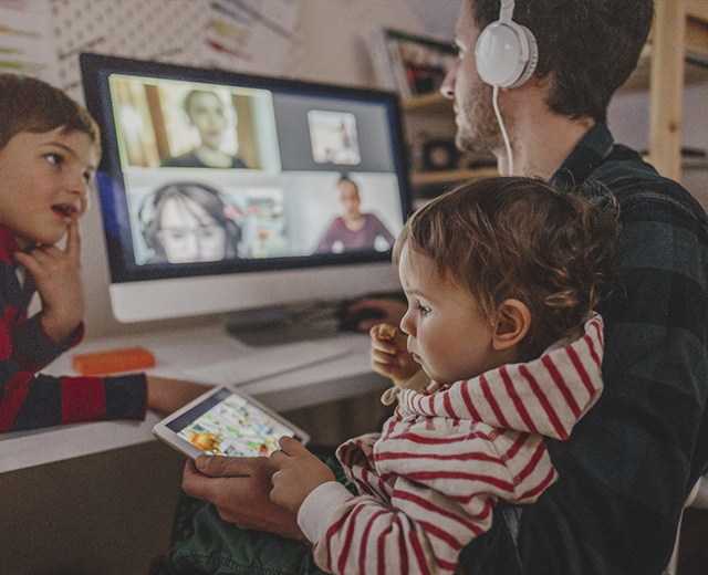 Man at home computer in a video meeting while wearing headphones with two children on his lap